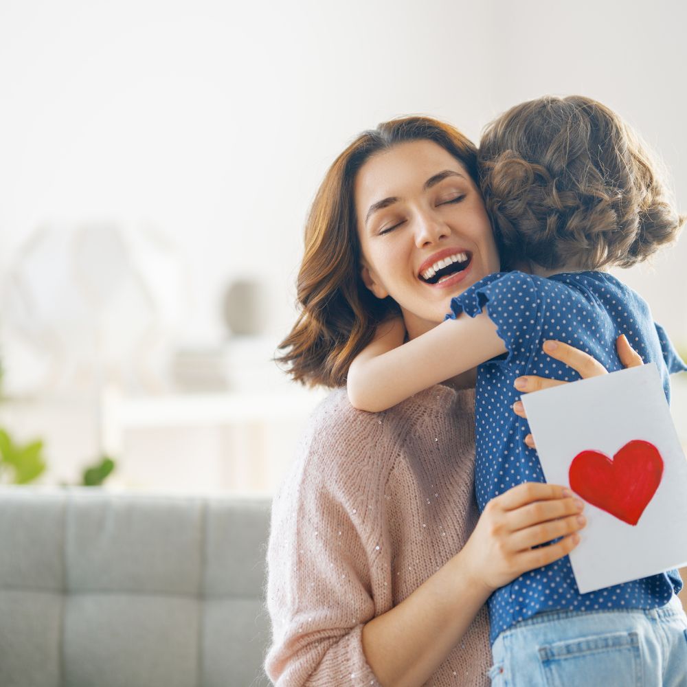 Mother smiling with eyes closed while hugging her young daughter who holds a handmade card with a red heart – emotional moment perfect for personalised Mother’s Day gift listings