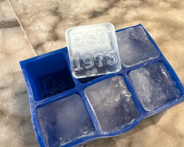 Five-star customer review photo showing a personalised 'EST 1975' ice cube from a blue silicone tray by Chilled Out Creations. The clear, raised lettering on the ice cube celebrates a special year, captured in a real home setting on a marble countertop.