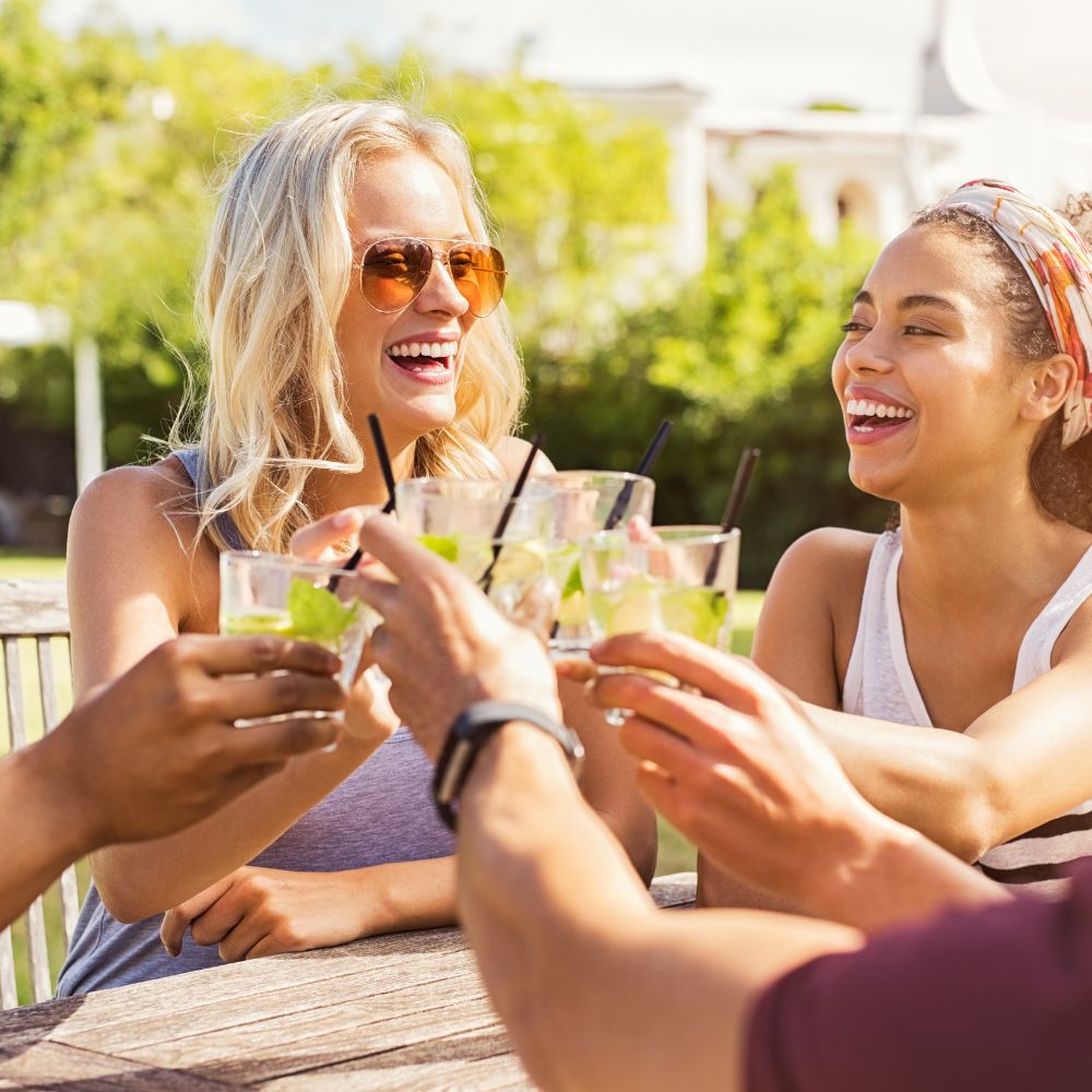 Group of smiling friends raising cocktail glasses outdoors on a sunny day, celebrating with a cocktail-loving mum in a fun and relaxed garden setting.