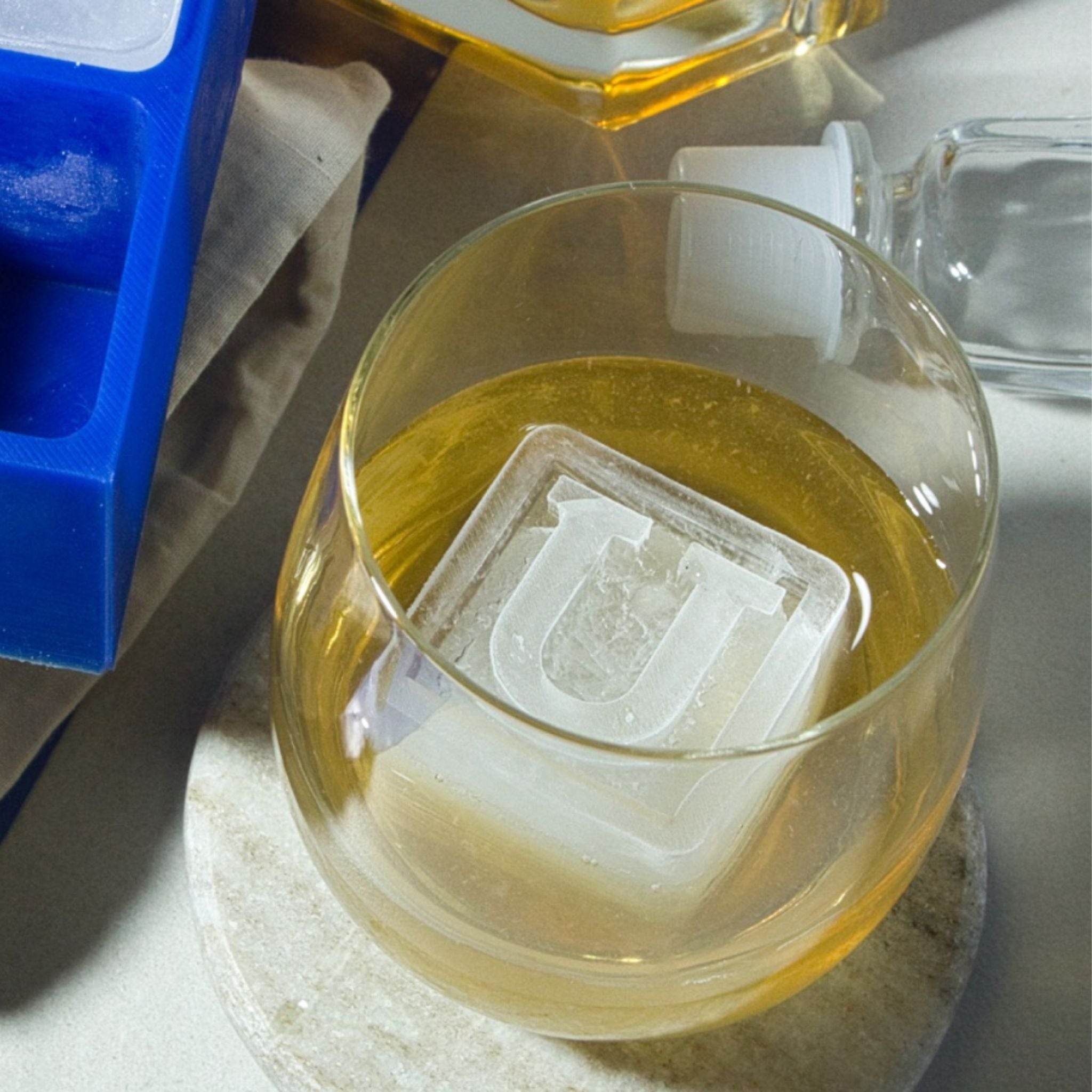 Close-up of a square ice cube with the letter U in a whiskey glass, next to a blue ice tray and bottle – perfect for unique milestone birthday gifts or personalised celebrations.