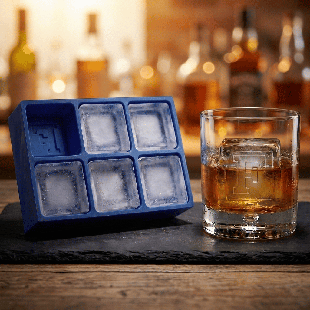 A blue silicone ice cube tray with six square compartments sits on a dark slate board next to a clear glass tumbler filled with an amber whiskey and a large ice cube, with blurred liquor bottles in the background.