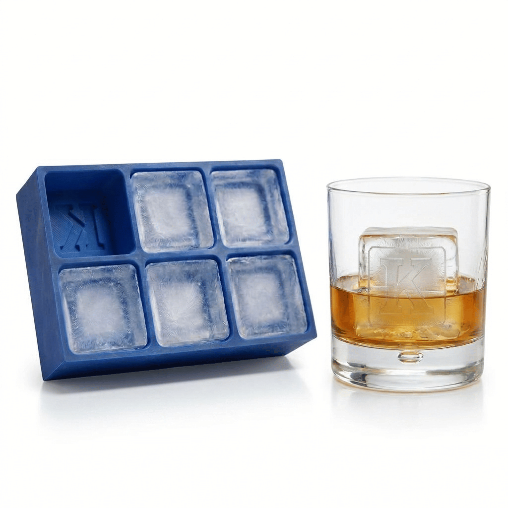 Blue silicone Letter K ice cube tray beside a rocks glass of amber whiskey chilled with a large clear ice cube stamped with the letter K, on a white background.