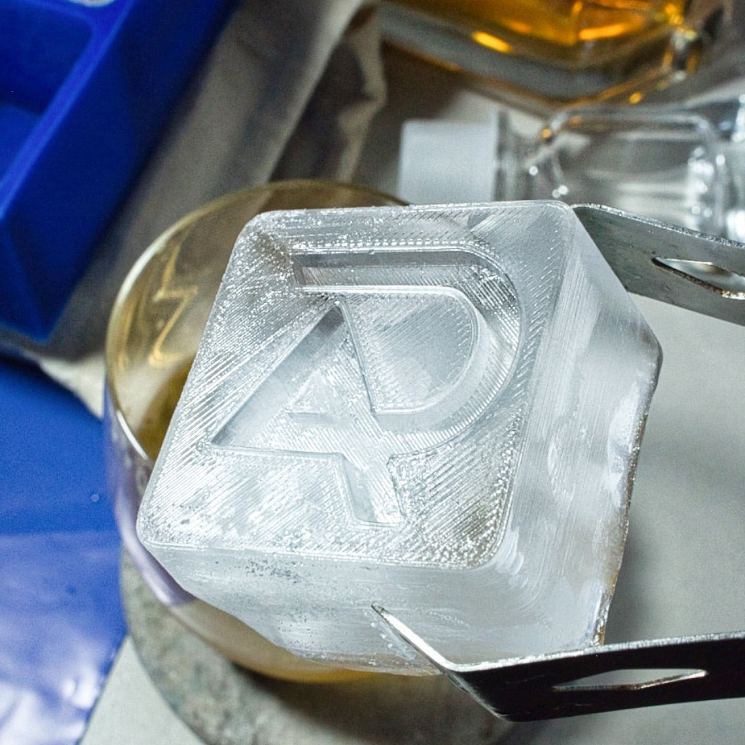Close-up of a large custom ice cube with engraved AP logo, held by tongs over a whiskey glass – perfect for business gifting or brand promotions.