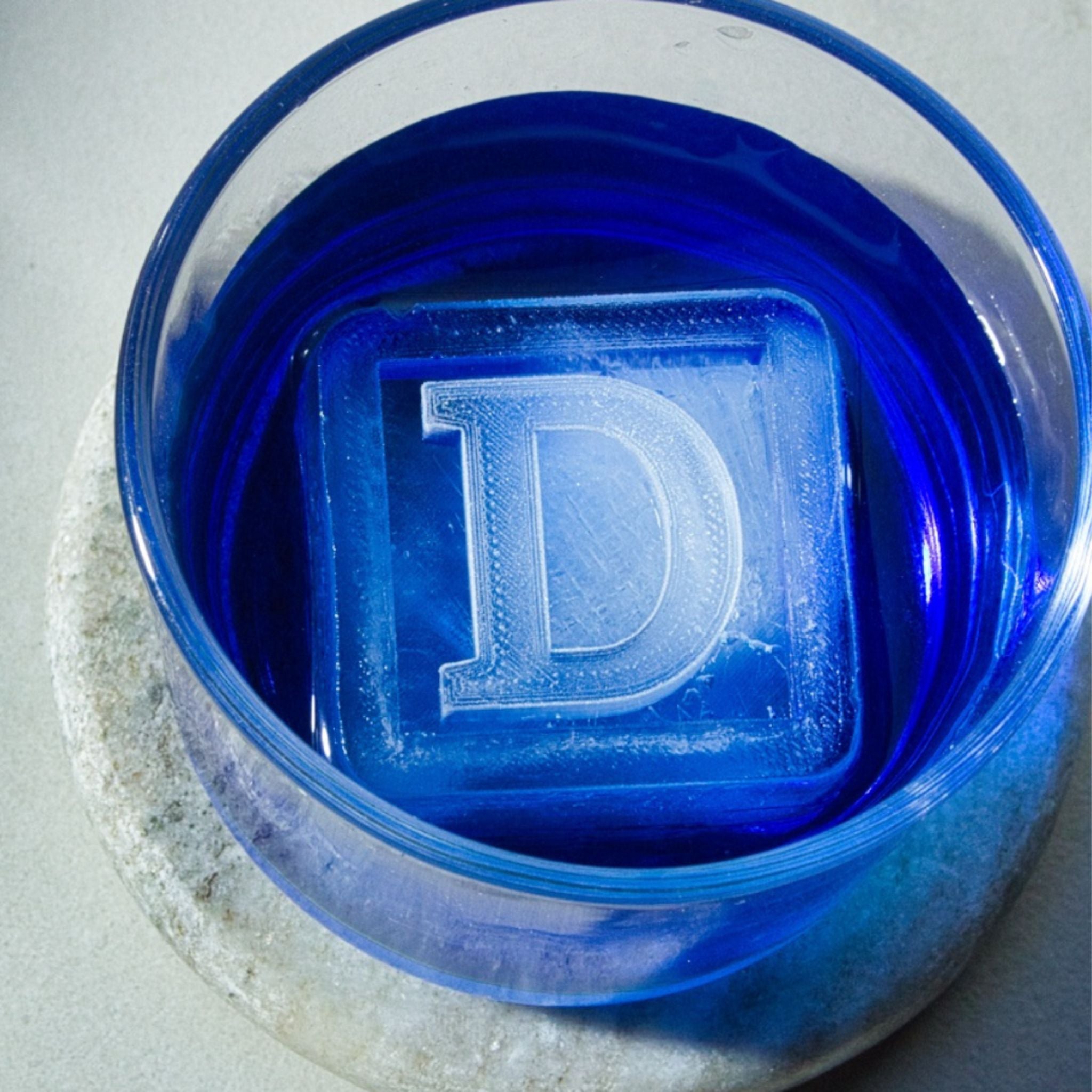 Clear ice cube with raised letter D in a deep blue whiskey glass, perfect for personalised gift sets and milestone drinks