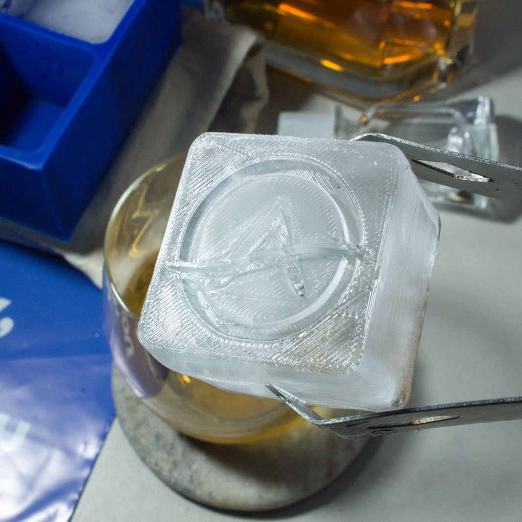 Close-up of a clear custom ice cube featuring a branded logo, held with tongs above a whiskey glass – perfect for business gifts and brand impressions.