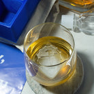 Whiskey glass holding a branded ice cube with gear and crossed tool design, set on bar coaster.