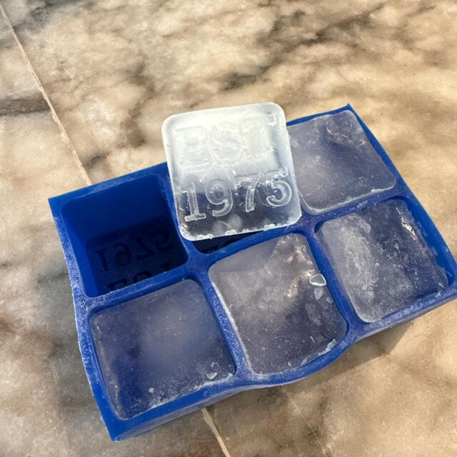 Five-star customer review photo showing a personalised 'EST 1975' ice cube from a blue silicone tray by Chilled Out Creations. The clear, raised lettering on the ice cube celebrates a special year, captured in a real home setting on a marble countertop.