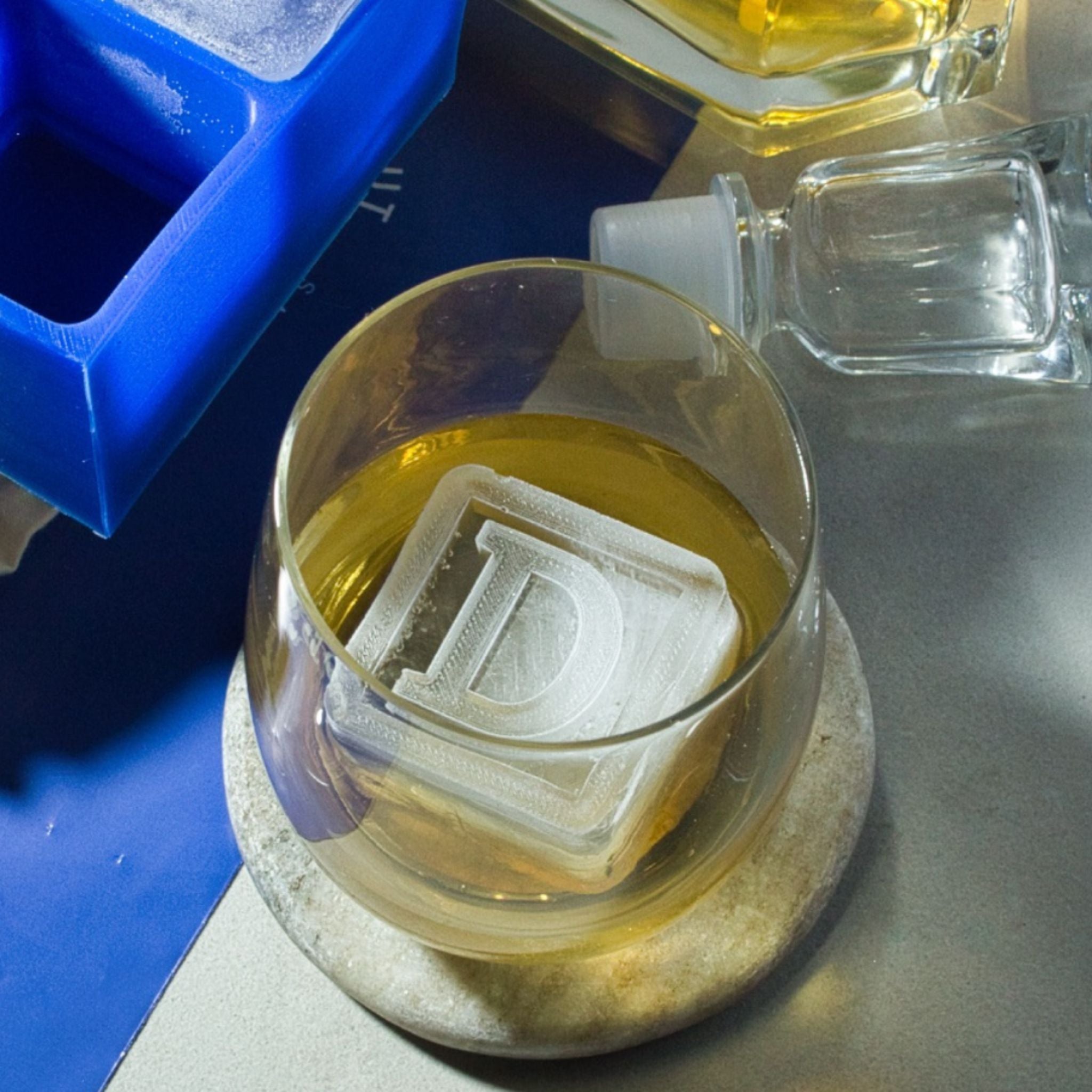 Raised D monogram ice cube in a whiskey glass with blue tray in the background, ideal for showcasing custom ice gifts