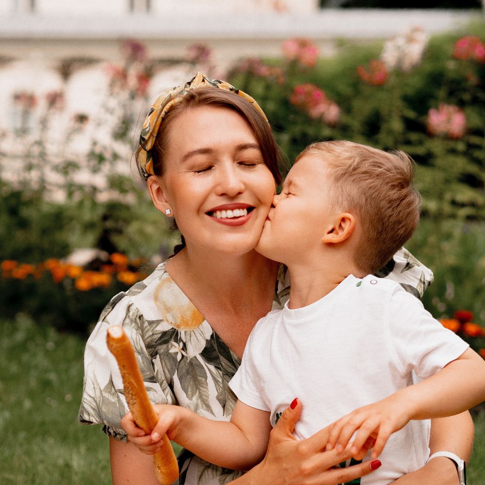 Young boy kisses smiling mum on the cheek while holding breadsticks in a flower-filled garden – heartfelt Mother’s Day moment that suits gifting ice trays from children