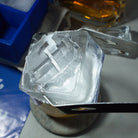A frosted ice cube with embossed gear and hammer design being held with metal tongs above whiskey glass.