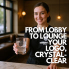 Smiling hotel staff member offers a rocks glass with a 45 mm crystal-clear ice cube embossed with the Velora Stay leaf logo; overlay text reads “From Lobby to Lounge — Your Logo, Crystal-Clear”; hospitality promotional branding image.