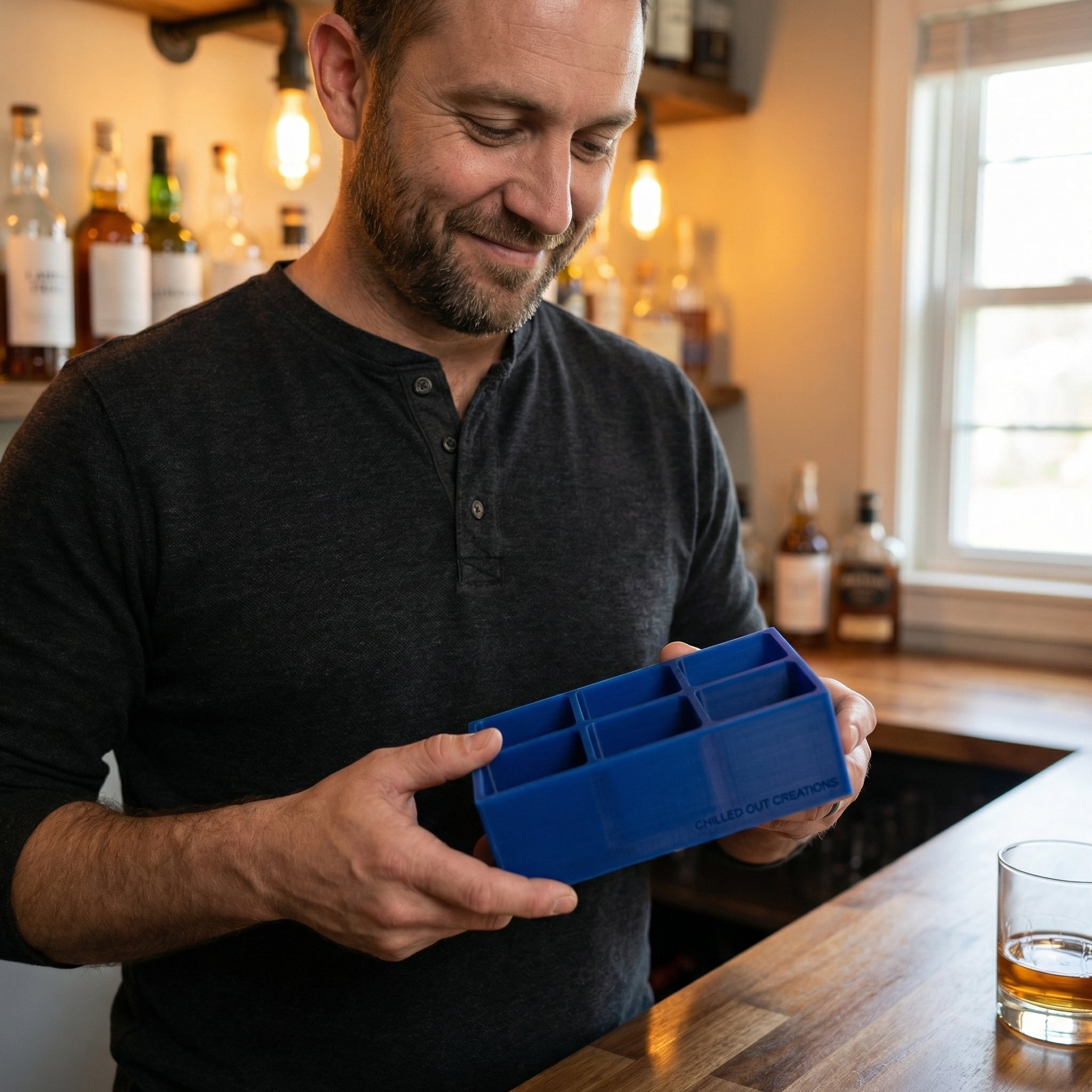Man at a home bar holding a blue silicone whiskey ice cube tray beside a glass of whiskey, a custom gift idea for him