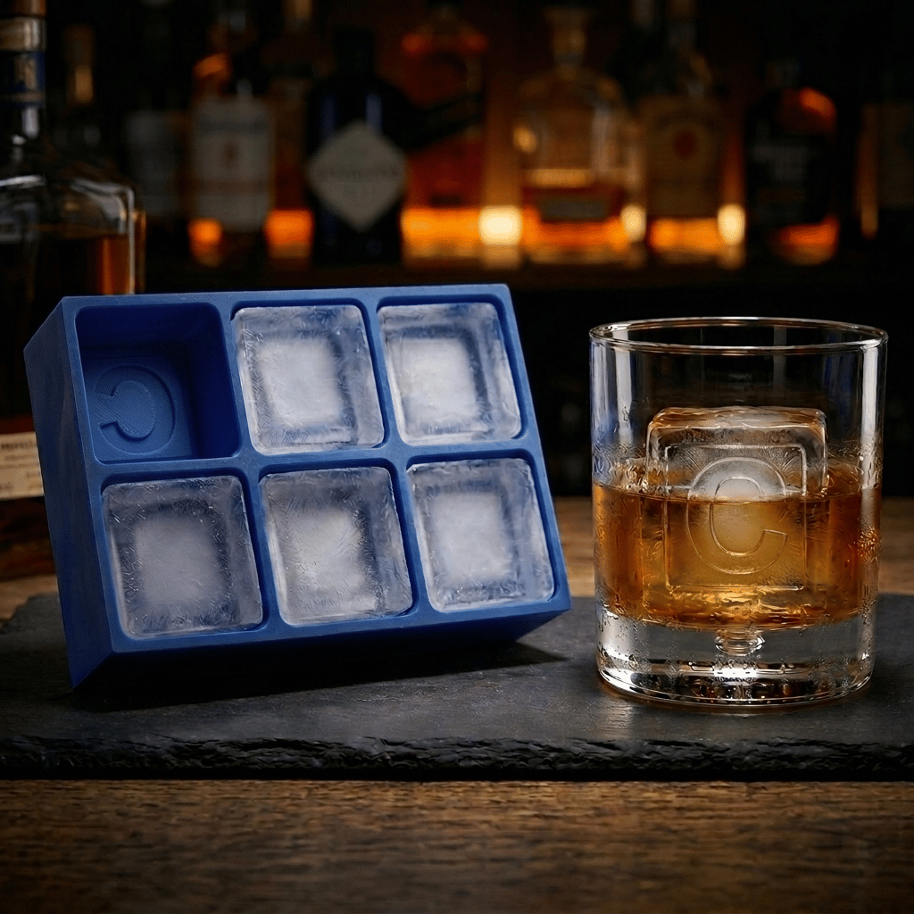 A blue silicone ice cube tray with six square compartments sits on a dark slate board next to a clear rocks glass filled with an amber drink, with blurred bar bottles in the background.