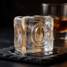 A clear square ice cube with a raised letter 'C' embossed on one side, resting on a dark slate surface with a blurred amber drink and dark background.