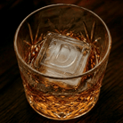 A cut crystal whiskey tumbler filled with amber whiskey and a large ice cube embossed with the letter D, set on a dark wooden surface.