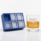 A blue silicone ice cube tray with six compartments sits next to a clear rocks glass filled with an amber drink and a large square ice cube, on a white background.