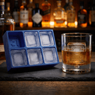 A blue silicone ice cube tray with six square compartments sits on a dark slate board next to a clear glass tumbler filled with an amber whiskey and a large ice cube, with blurred liquor bottles in the background.
