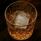 A cut-crystal whiskey glass filled with amber liquor and a large clear ice cube stamped with the letter E, set on a dark wooden surface.