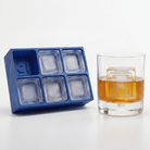 A blue silicone ice cube tray with six square compartments sits next to a clear rocks glass filled with an amber drink and a large square ice cube, on a clean white background.