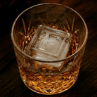 A cut-crystal whiskey glass filled with amber liquor and a large clear ice cube stamped with the letter F, set on a dark wooden surface.