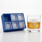 A blue silicone ice cube tray with six compartments sits next to a clear glass tumbler filled with an amber drink on a white studio background.