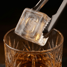 Stainless steel ice tongs gripping a large clear ice cube stamped with the letter G above a cut-crystal glass of amber whiskey.