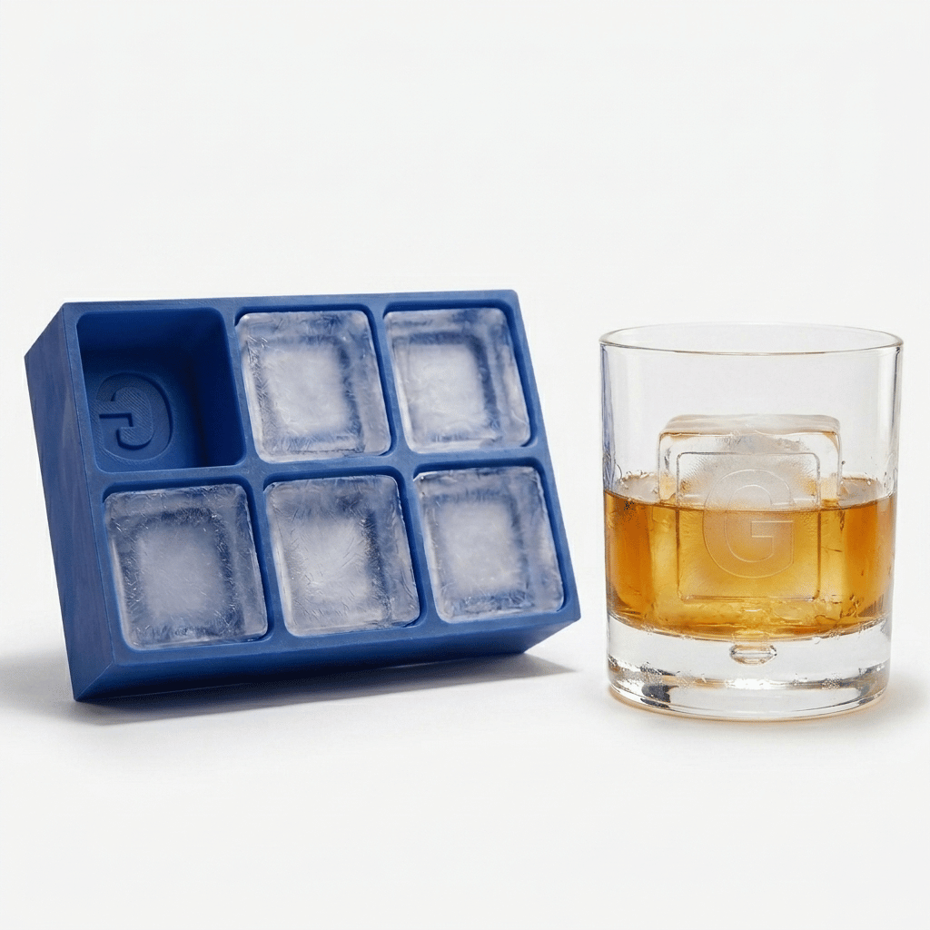 Blue silicone Letter G ice cube tray beside a rocks glass of amber whiskey chilled with a large clear ice cube stamped with the letter G, on a white background.