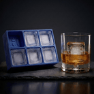 Blue silicone Letter H ice cube tray and a rocks glass of amber whiskey with a large H-stamped ice cube, on a dark slate surface against a moody black background.