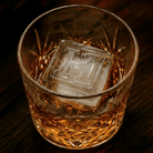 A cut-crystal whiskey glass filled with amber liquor and a large clear ice cube stamped with the letter H, set on a dark wooden surface.