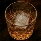 A cut-crystal whiskey glass filled with amber liquor and a large clear ice cube stamped with the letter I, set on a dark wooden surface.