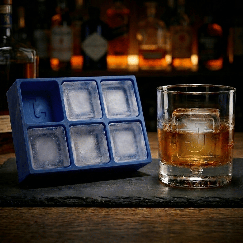 Blue silicone Letter J ice cube tray and a whiskey glass with a J-stamped ice cube on a slate board, set against a warmly lit back-bar of liquor bottles.