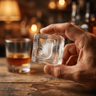 A person's hand holding up a large clear ice cube stamped with the letter J, with a glass of amber whiskey and a warmly lit bar in the background.