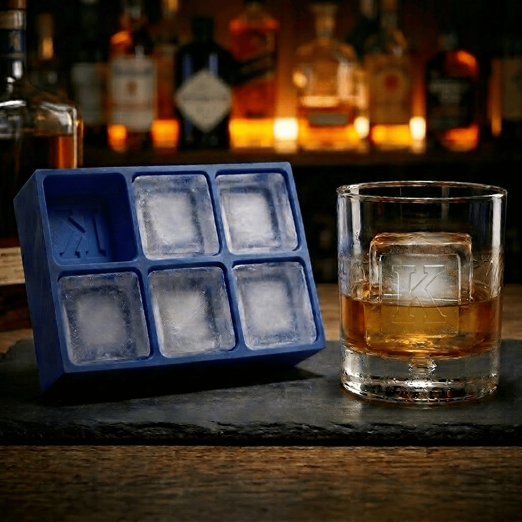 Blue silicone Letter K ice cube tray and a whiskey glass with a K-stamped ice cube on a slate board, set against a warmly lit back-bar of liquor bottles.