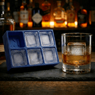 Blue silicone Letter K ice cube tray and a whiskey glass with a K-stamped ice cube on a slate board, set against a warmly lit back-bar of liquor bottles.