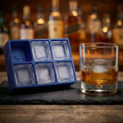 Blue silicone Letter L ice cube tray and a whiskey glass with an L-stamped ice cube on a slate board, set against a warmly lit back-bar of liquor bottles.