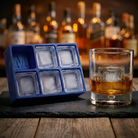 Blue silicone Letter M ice cube tray and a whiskey glass with an M-stamped ice cube on a wooden bar top, set against a warmly lit back-bar of liquor bottles.