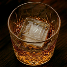 A cut-crystal whiskey glass filled with amber liquor and a large clear ice cube stamped with the letter M, set on a dark wooden surface.