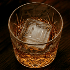 A cut-crystal whiskey glass filled with amber liquor and a large clear ice cube stamped with the letter N, set on a dark wooden surface.