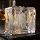A large clear ice cube stamped with the letter N resting on a black slate coaster, with a glass of amber whiskey blurred in the background.