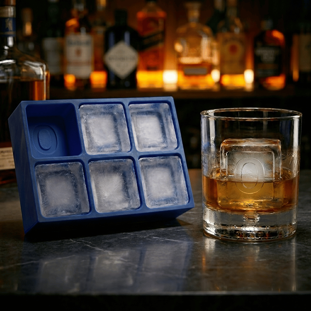Blue silicone Letter O ice cube tray and a whiskey glass with an O-stamped ice cube on a dark stone bar top, set against a warmly lit back-bar of liquor bottles.