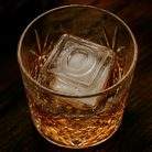 A cut-crystal whiskey glass filled with amber liquor and a large clear ice cube stamped with the letter O, set on a dark wooden surface.
