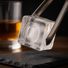 Stainless steel ice tongs holding a large clear ice cube stamped with the letter O above a black slate coaster, with a glass of amber whiskey in the background.