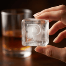 A person's hand holding up a large clear ice cube stamped with the letter P, with a glass of amber whiskey blurred in the background.