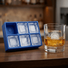 Blue silicone Letter Q ice cube tray and a whiskey glass with a Q-stamped ice cube on a wooden bar top, set against a warmly lit back-bar.