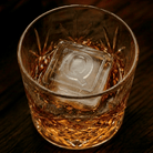 A cut-crystal whiskey glass filled with amber liquor and a large clear ice cube stamped with the letter Q, set on a dark wooden surface.