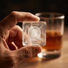A person's hand holding up a large clear ice cube stamped with the letter Q, with a glass of amber whiskey blurred in the background.