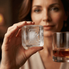 A woman holds a large clear ice cube stamped with the letter R up to the camera, with a glass of whiskey in the background.