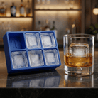 A blue silicone Letter T ice tray beside a whiskey glass holding a clear ice cube stamped with the letter T, set on a dark bar counter with backlit bottles behind.
