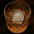 A cut-crystal whiskey glass filled with amber liquor and a large clear ice cube stamped with the letter T, set on a dark wooden surface.