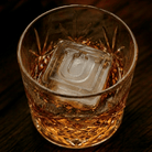 A cut-crystal whiskey glass filled with amber liquor and a large clear ice cube stamped with the letter U, set on a dark wooden surface.